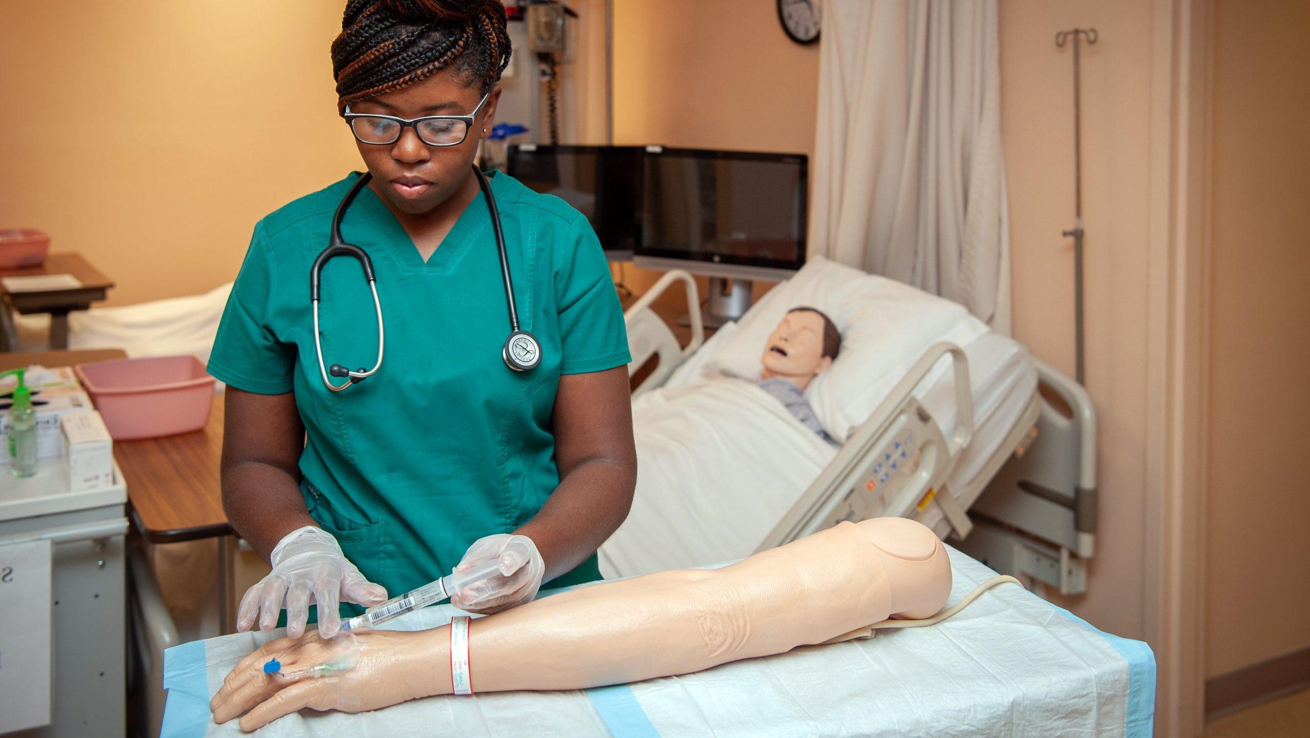 Female student in nursing simulation lab using syringe and tube on mannequin arm.